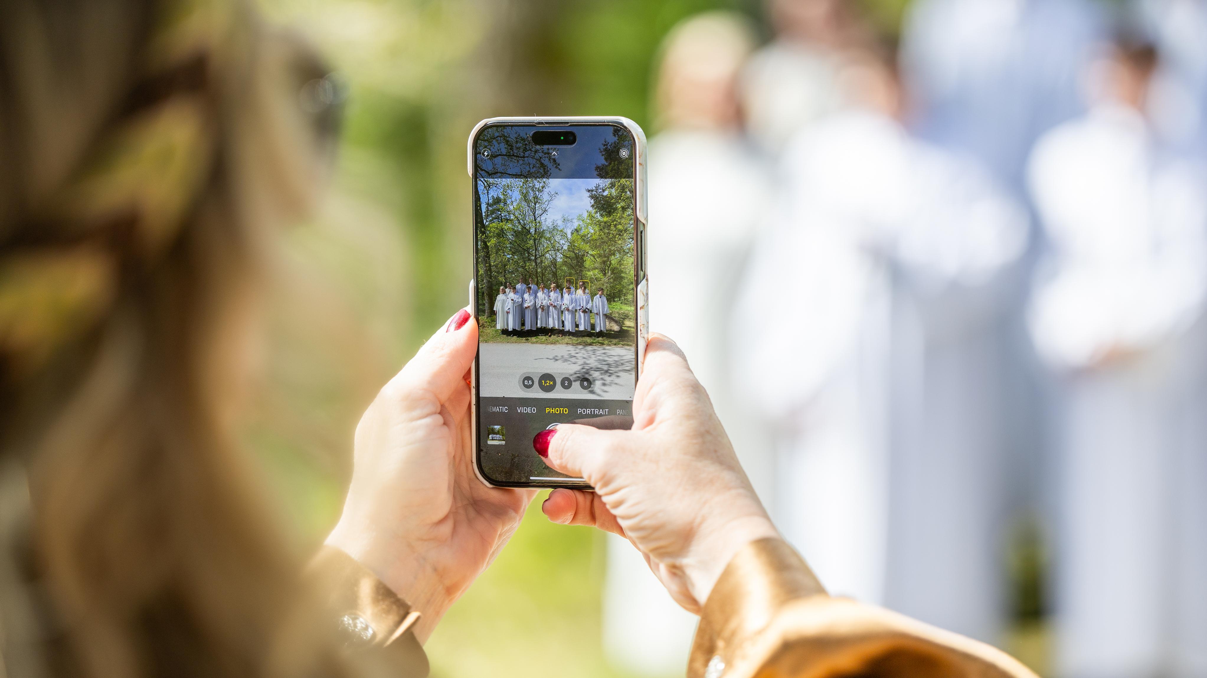 en kvinne som fotograferer en gjeng konfirmanter i hvite konfirmantkapper med mobilen