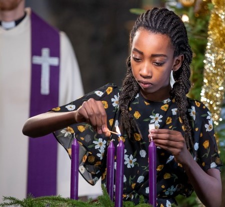 Ei jente tenner adventslysene. I bakgrunnen står presten, og vi ser et pyntet juletre.