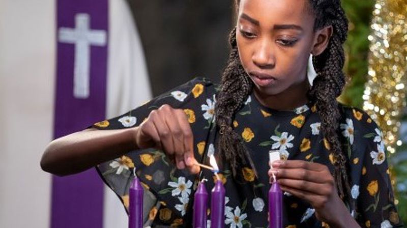 Ei jente tenner adventslysene. I bakgrunnen står presten, og vi ser et pyntet juletre.