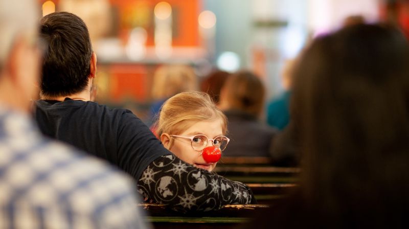 Jente sitter med rød klovnenese i armkroken til en voksen