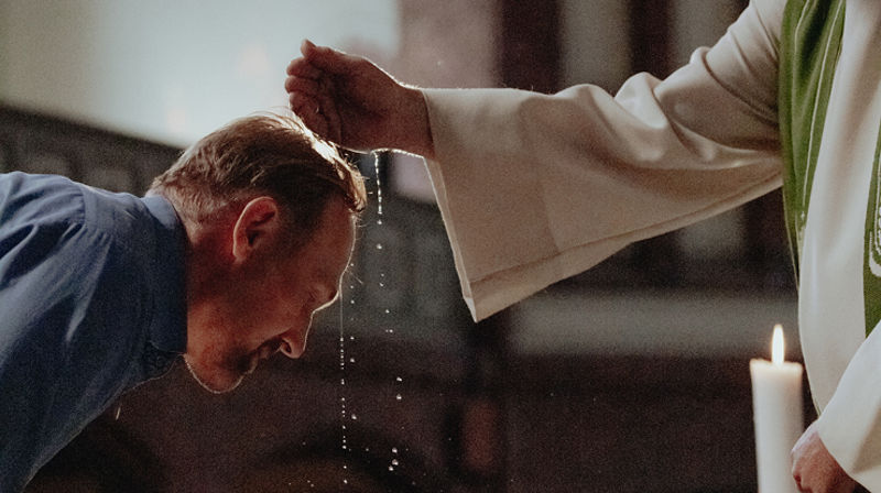 En eldre mann lener seg over døpefonten og lar seg døpe.