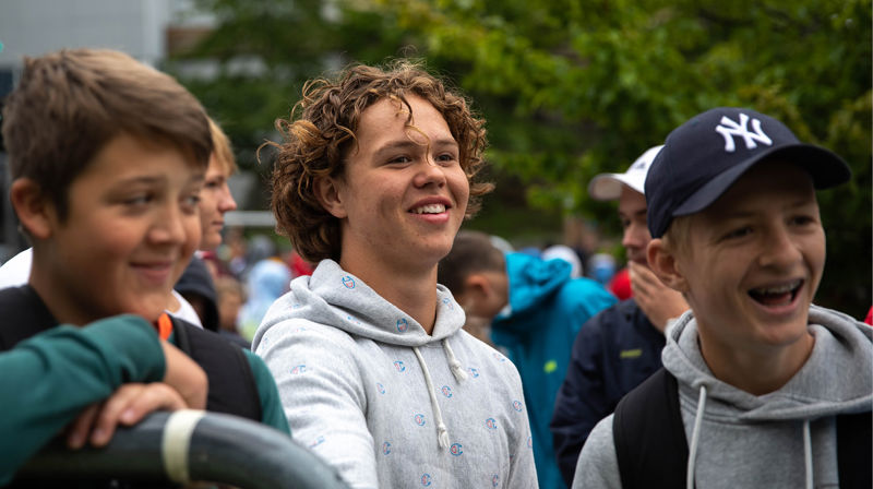 Tre gutter i en folkemengde. De følger interessert med på noe og smiler. Det er sommer og konfleir.