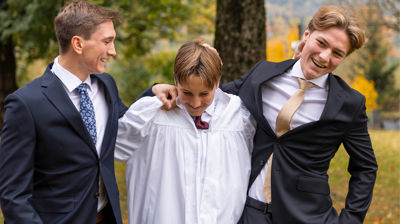 Tre gutter, en av dem har på konfirmantkappe, de to andre har på dress. De ler.