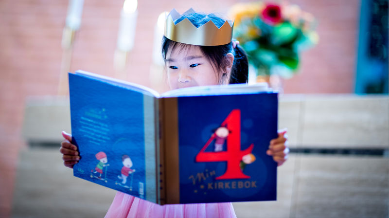 Jente med krone på hodet leser i 4årsboka. Hun står foran alteret i et kirkerom. På alteret står det blomster.