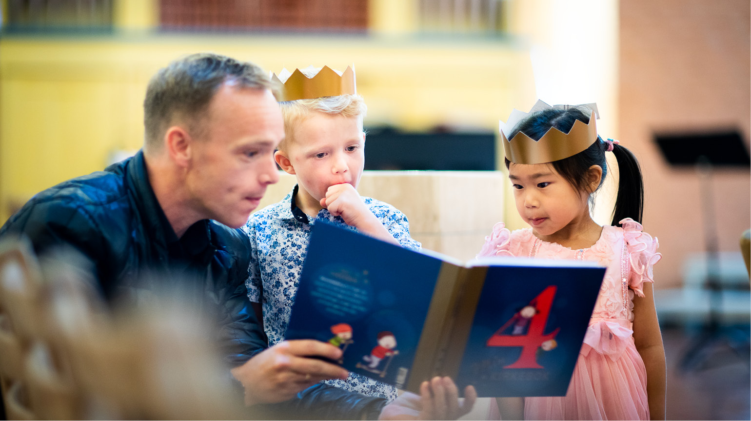 En mann ser i 4årsbok sammen med to barn, Begge barna har fått krone på hodet. De kikker interesserte på boka. 