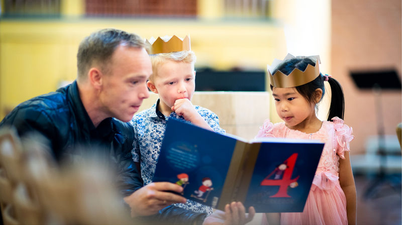 En mann ser i 4årsbok sammen med to barn, Begge barna har fått krone på hodet. De kikker interesserte på boka.