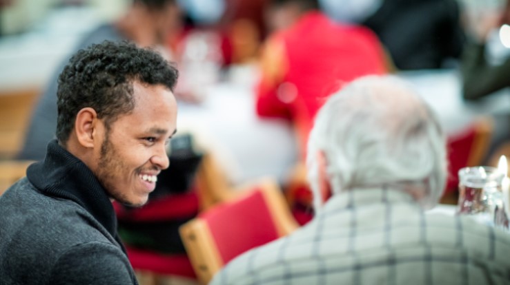 En ung mann smiler og har en god samtale med en eldre mann. Språkkafé og kirkekaffe er flotte fellesskap i kirken.