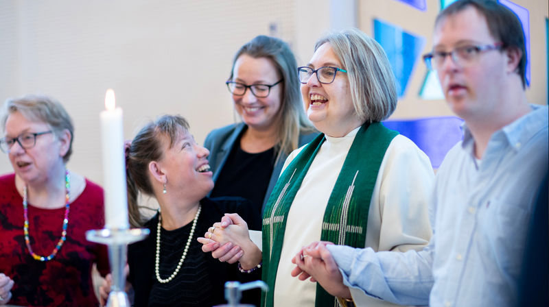 En gruppe mennesker holder hverandre i hendene, smiler og fremfører noe foran menigheten