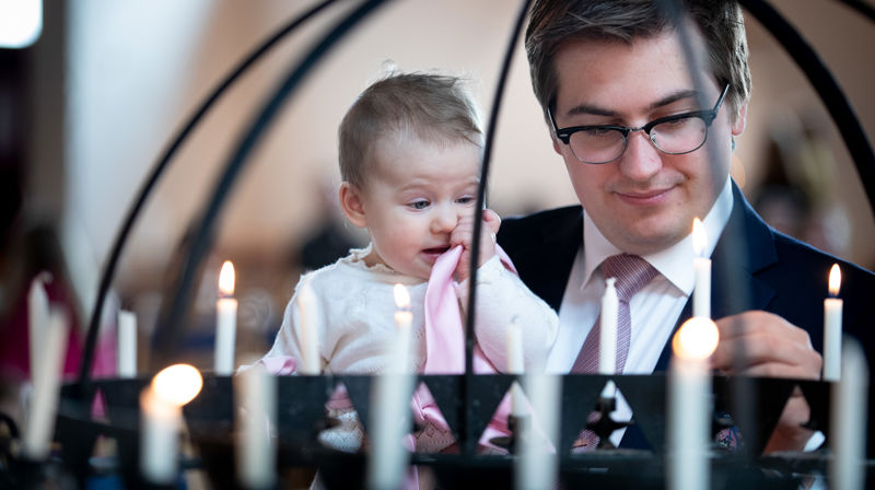 En far tenner et lys i lysgloben. Han bærer dåpsbarnet på armen. De smiler.