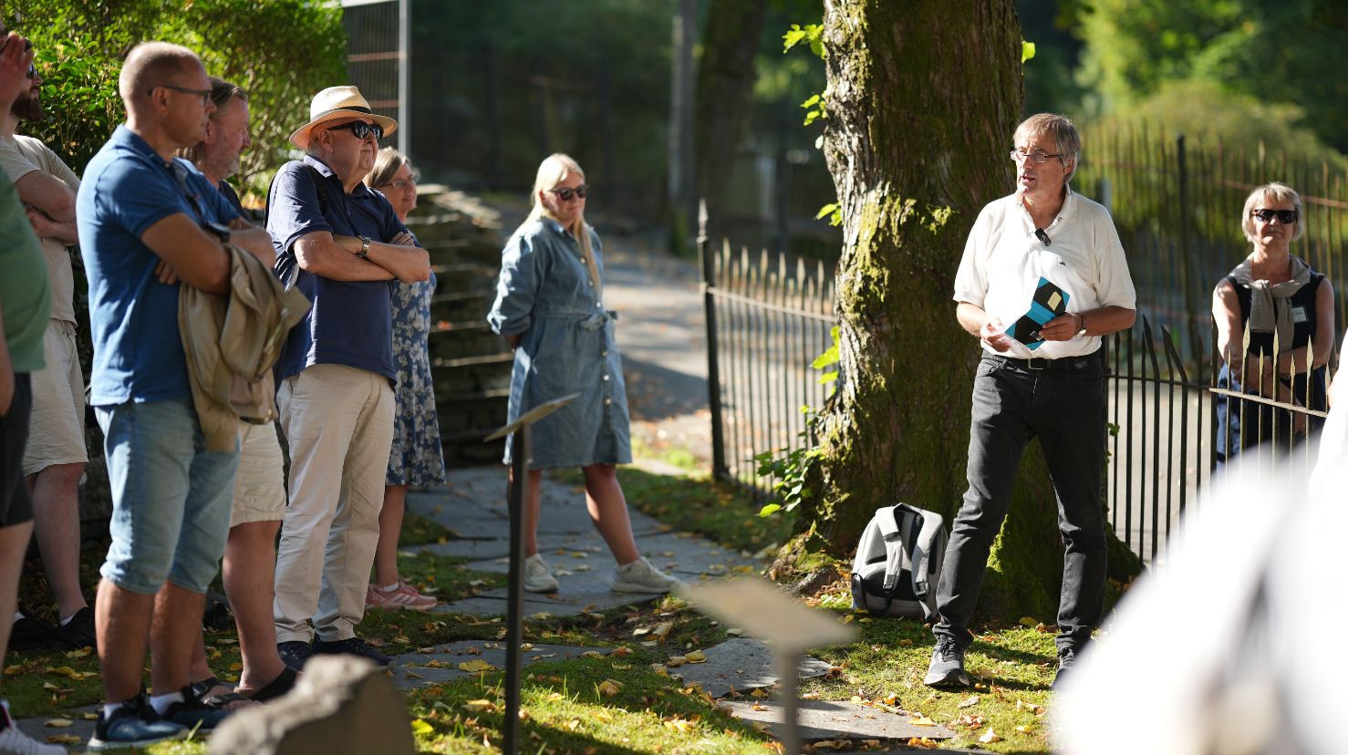 Mann snakker til folk som står på en gravplass i sol. Foto.