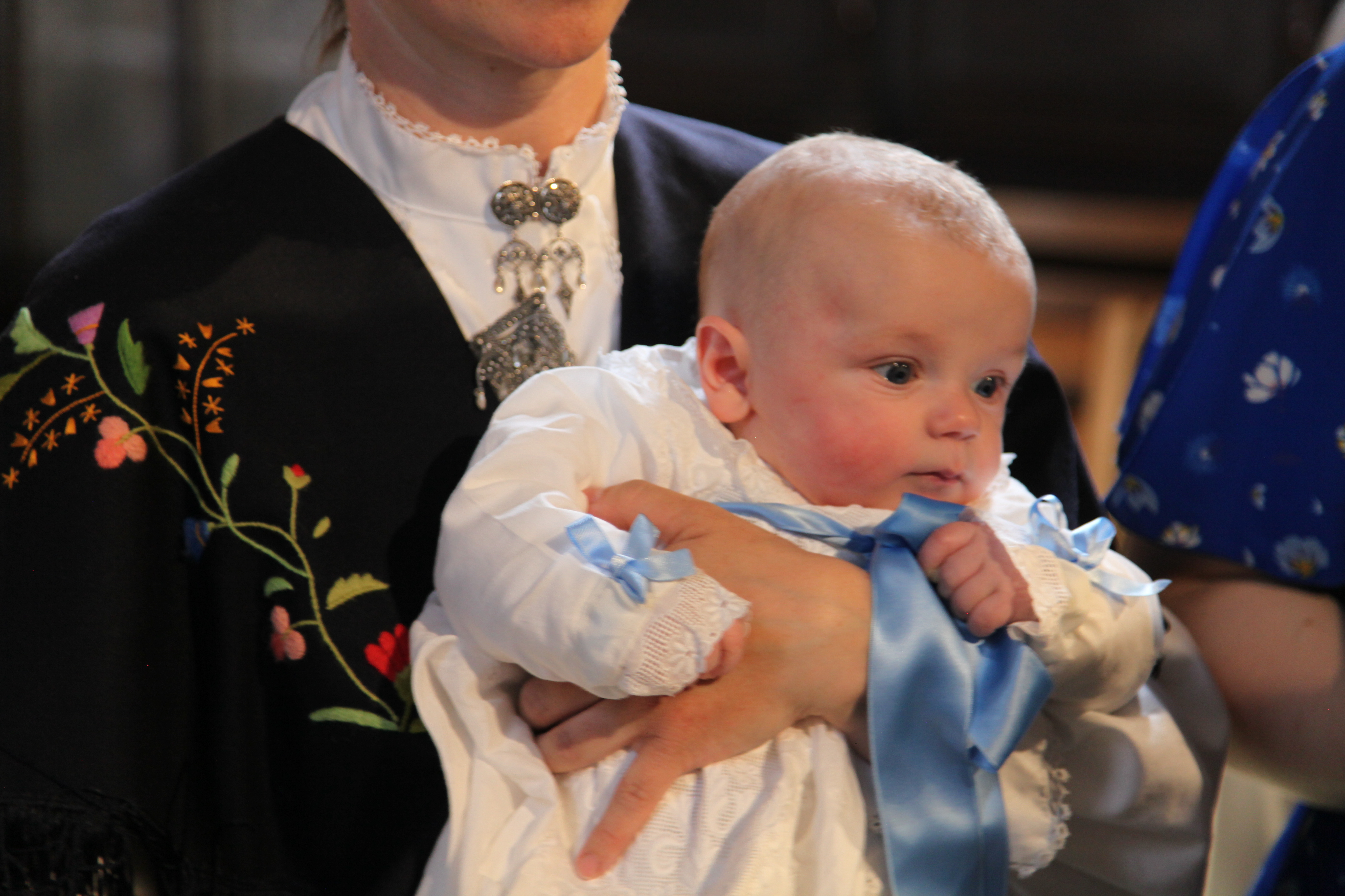 Dåpsbarn i hvit kjole holdt av forelder.