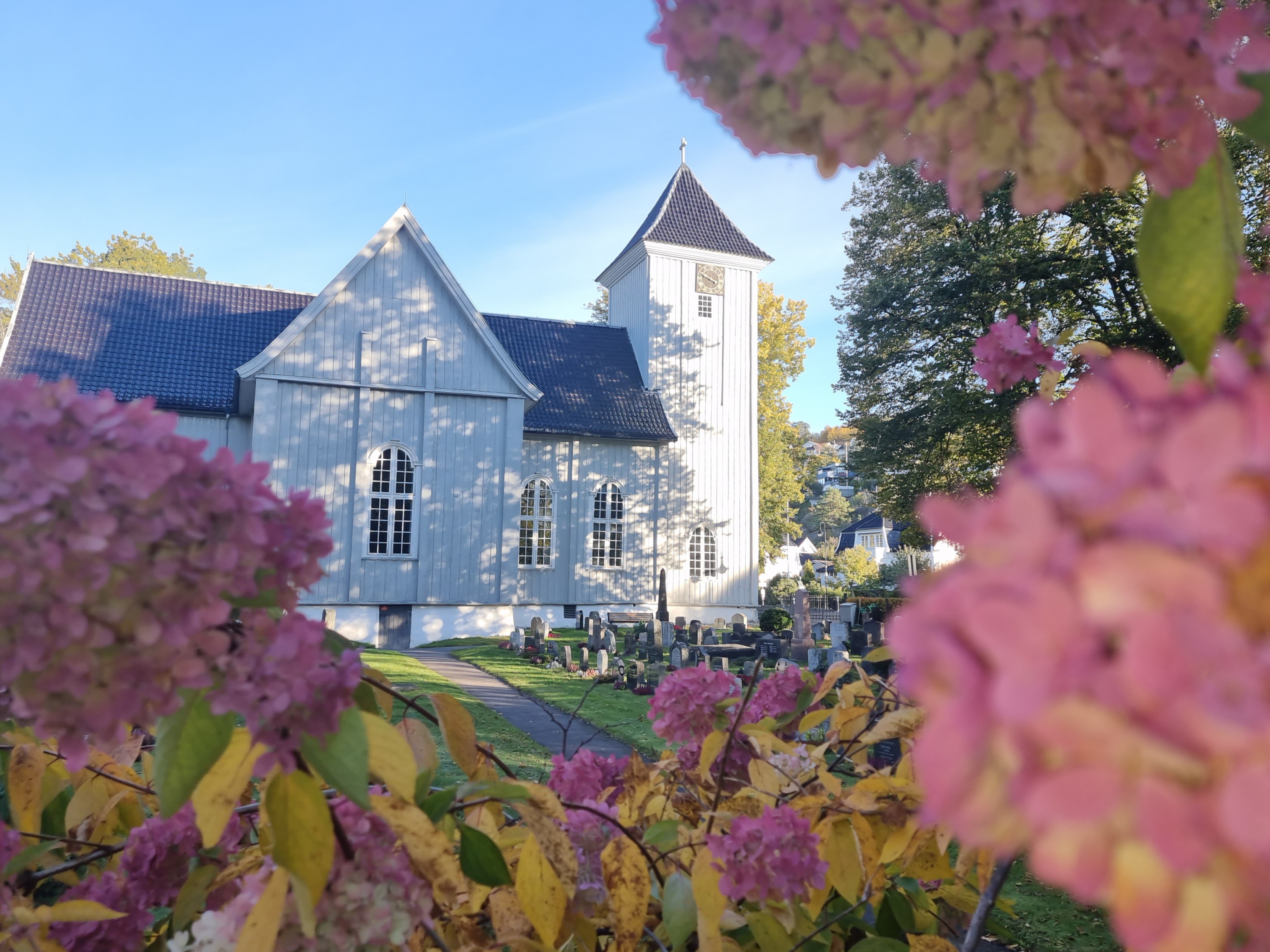 Kirken bader i sol og skygge fra trærne, rammet inn av blomster fra kirkegården.