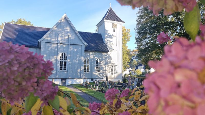 Kirken bader i sol og skygge fra trærne, rammet inn av blomster fra kirkegården.