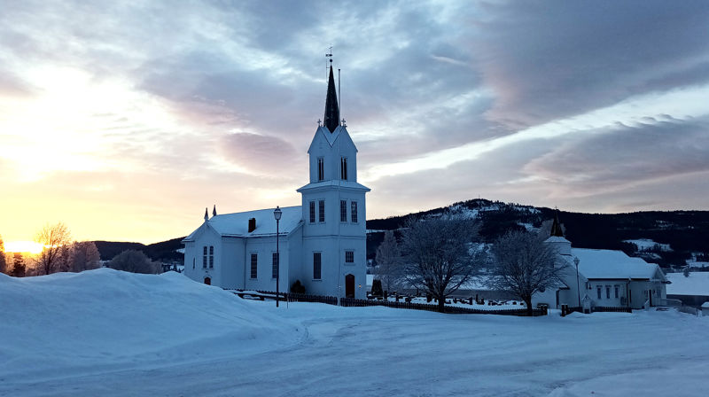 Olberg kirke og kirkestua dekket av snø mot en bakgrunn med skyer og sola som skinner gjennom.