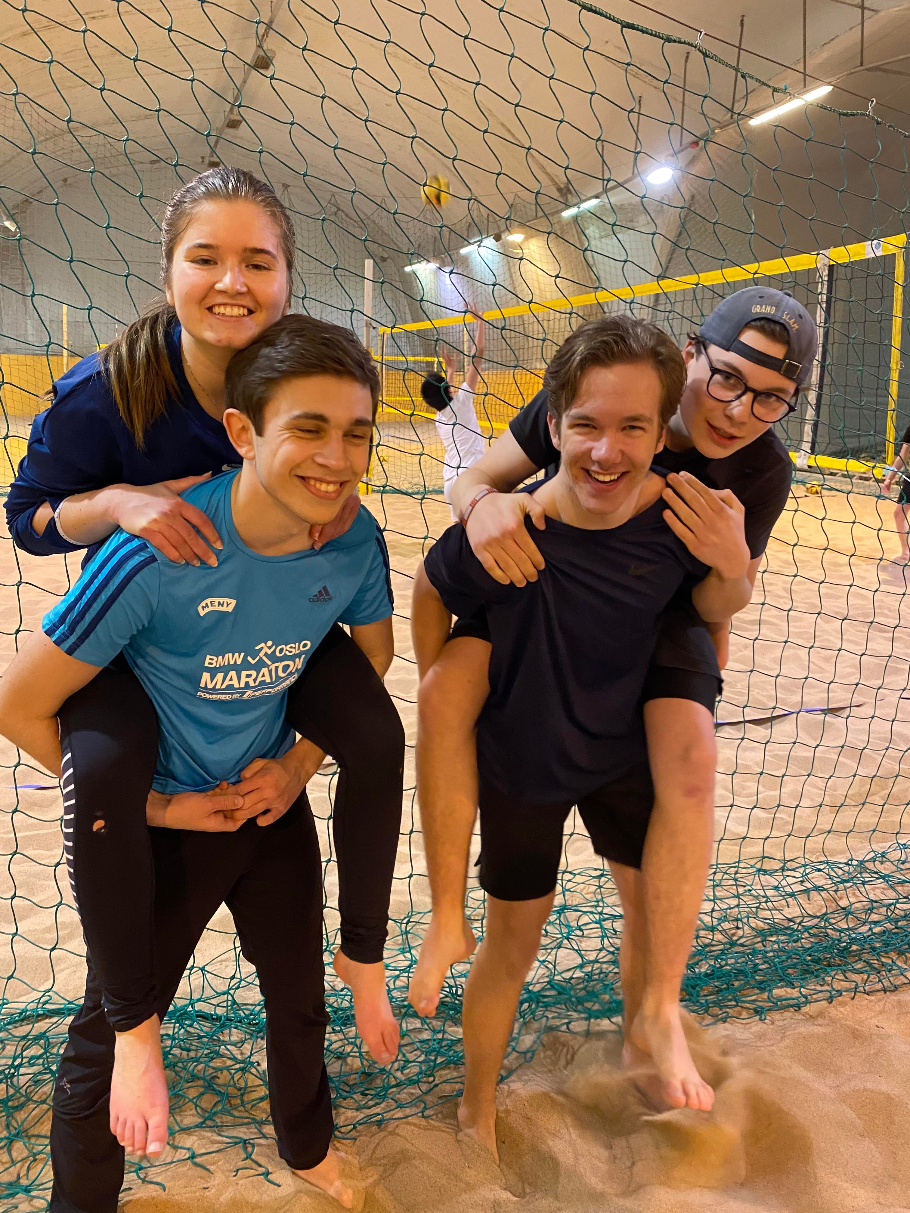 Bilde av to par ungdommer hvor hver av parene sitter på ryggen til hverandre og smiler stort. De står innendørs ved et volleyballnett