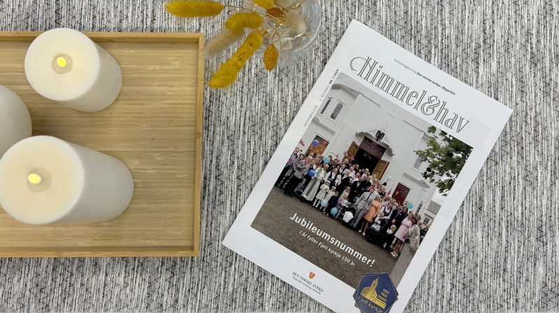 Et magasin med tittel "Himmel&hav" ligger på en grå duk på et bord. Ved siden av er et brett med tre elektriske lys, og en vase med pyntestrå.