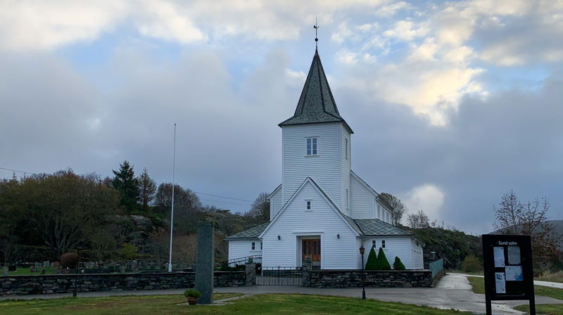 Kausland kyrkje med grå og blå himmel bak. Framfor er det grønt gras.