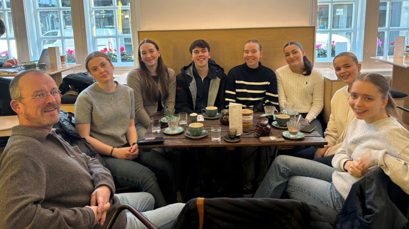 Sju ungdommer og en mann sitter rundt et bord på en kafe, de smiler mot kameraet. På bordet står ulike kopper og glass.