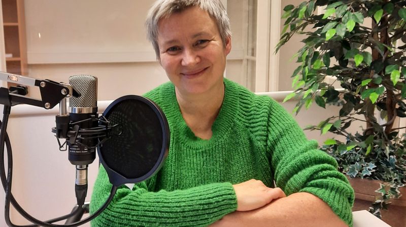 Sølvi helen sitter ved et bord med en mikrofon, hun lener seg på bordet og smiler