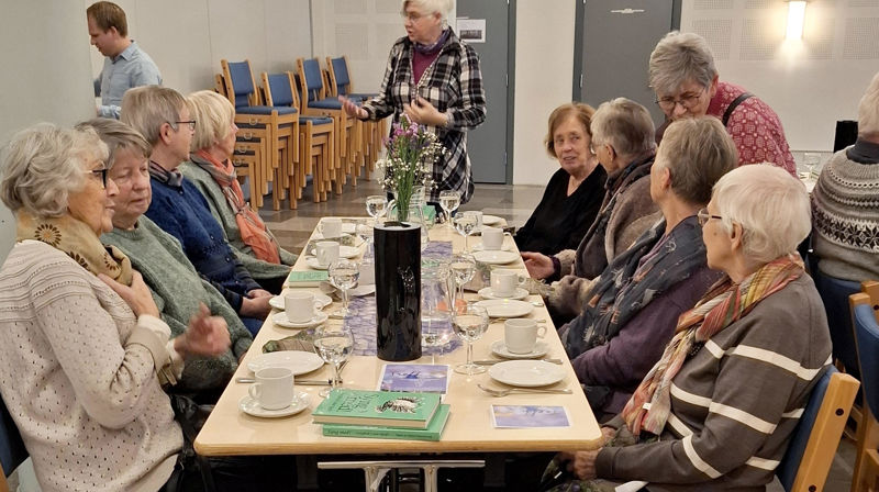 Flere eldre sitter ved et dekket bord, rundt står noen og snakker med de som sitter