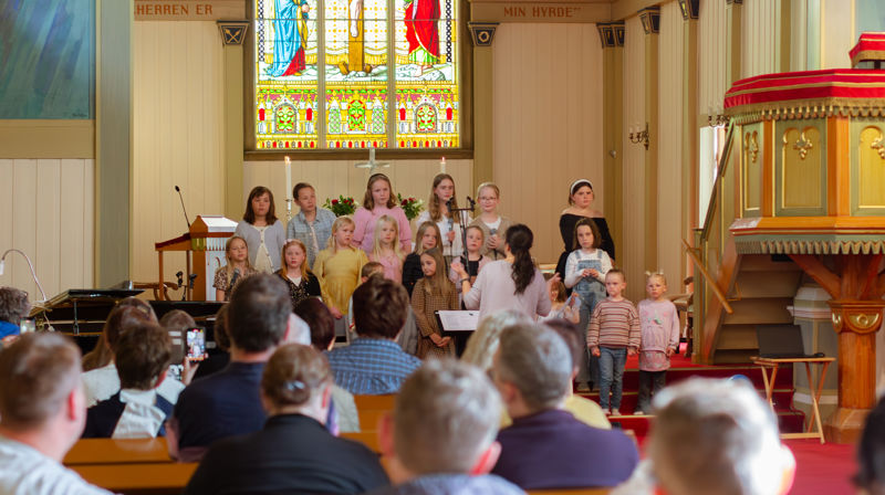 et barnekor står fremme og opptrer, i forgrunnen ser vi mennesker sitter i benkradene