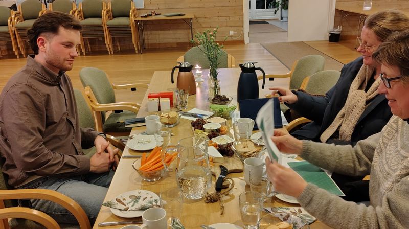 En mann og to damer sitter ved et bord og ser på noen ark. Bordet er fyllt med kjeks, ost, grønnsaker og dipp.