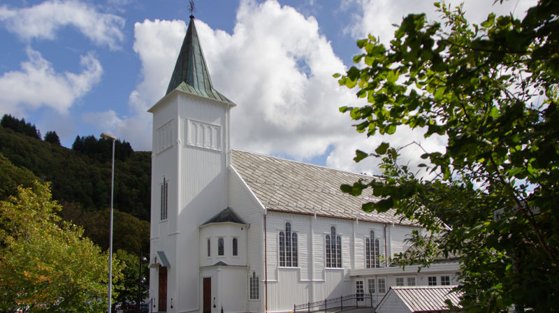 Fjell kyrkje i solskinn, det er blader på et tre på høyre side av bildet, treet skjuler kyrkjelydssalen.
