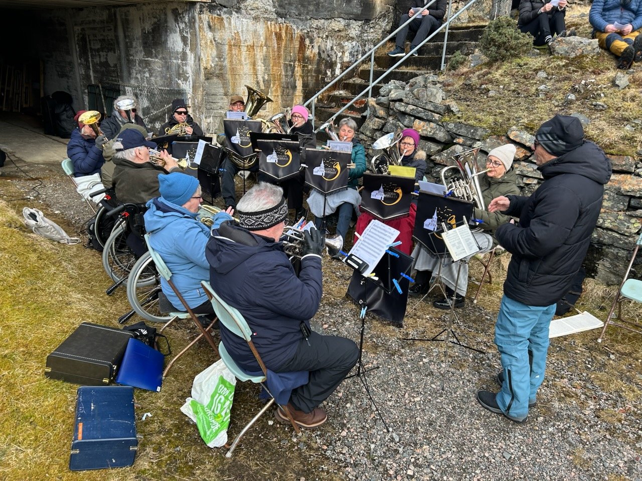 Et brassband sitter p&aring; stoler i en halvsirkel, de spiller ute og er godt kledd