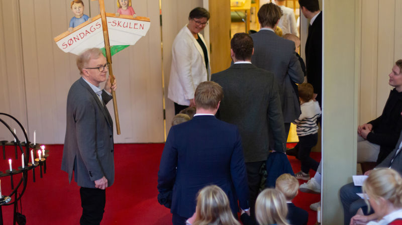En mann holder et skilt, på skiltet er det en båt med to barn og "søndagsskule" skrevet under de. Barn og voksne er på vei ut av kirkerommet.