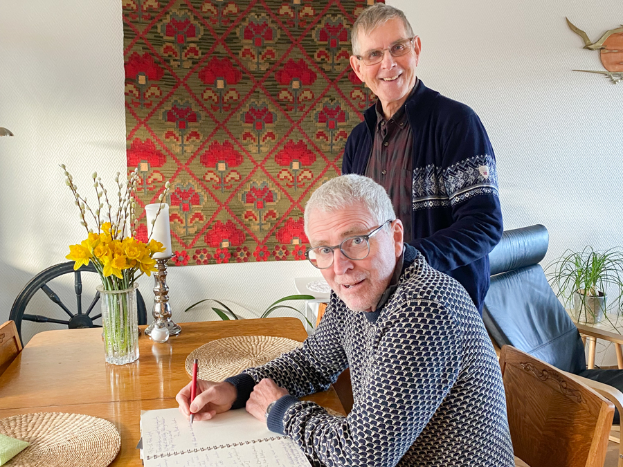 en mann sitter ved et bord og skriver, en annen st&aring;r ved siden av, begge ser mot kameraet