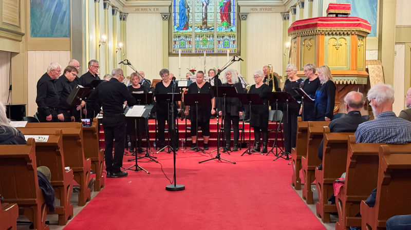 Koret står fremme i kirken i en halvsirkel og synger, vi kan se at mennesker sitter i benkradene