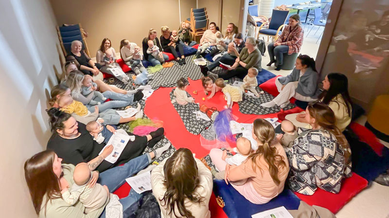 babyer og mammaer sitter inne i et lite rom i en stor sirkel, i midten er det tepper der noen babyer kryper