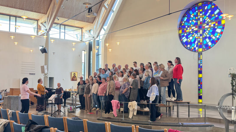 Et kor med rund 50 mennesker står framme bak alterringen og øver, en dirigent står til høyre i bildet sammen med to solister.