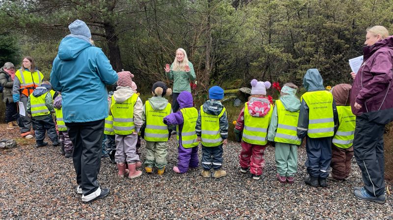 Ute på en grusvei står barnehagebarn med refleksvester i en halvsirkel, framme står en dame og forteller noe