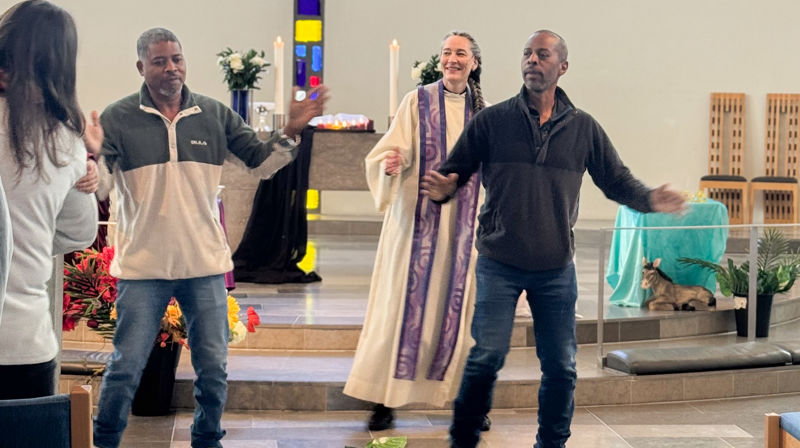 En kvinnelig prest og to menn står fremme i kirken og danser, mennene står fremst og kvinnen litt bak de.