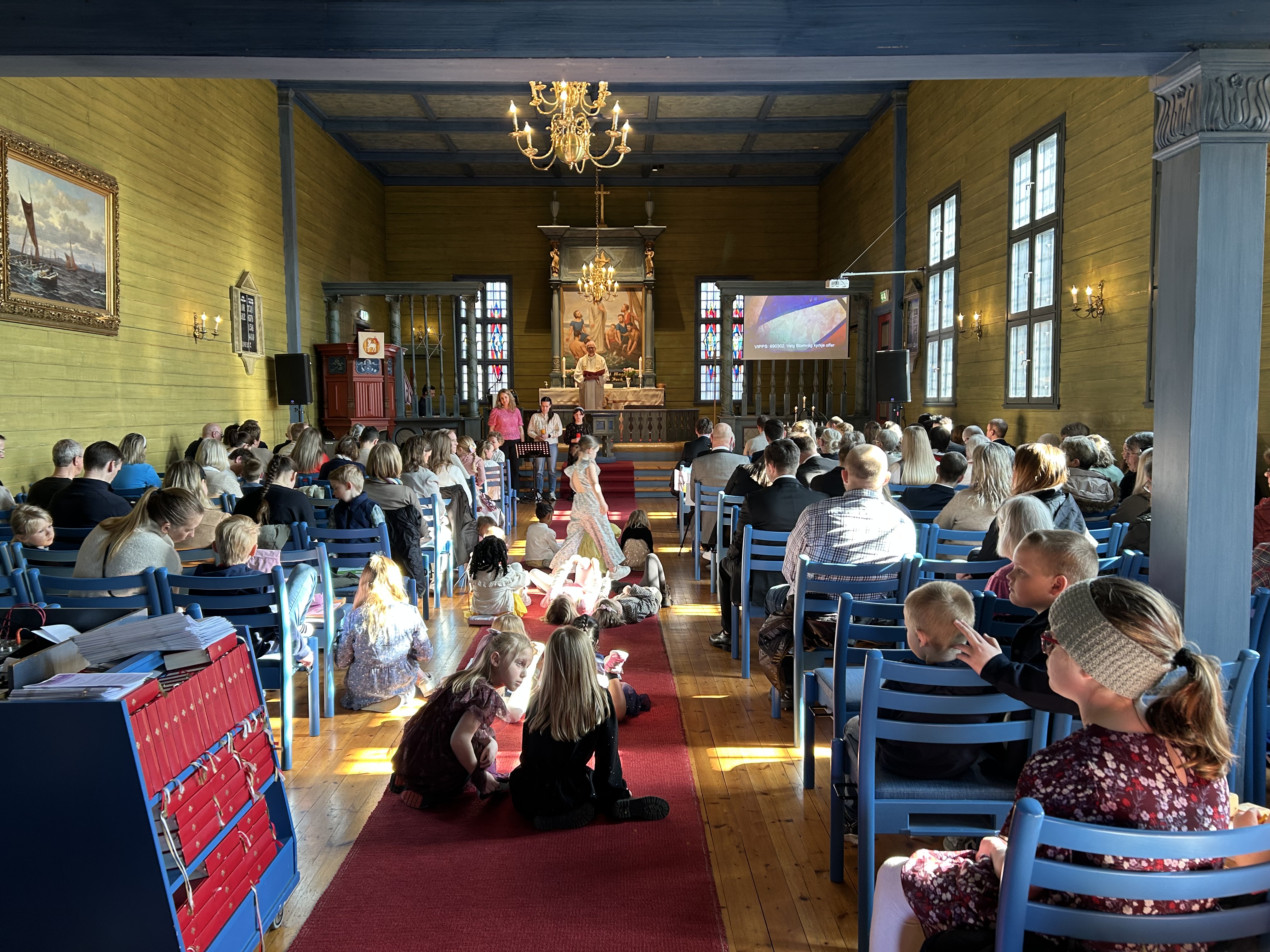 Det er fullt i kirken under en gudsteneste, barn sitter, står og ligger på teppet i midtgangen. fremme står tre forsangere, og presten står inne i alterringen.