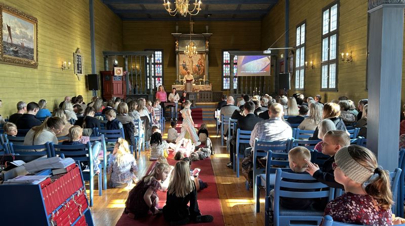 Det er fullt i kirken under en gudsteneste, barn sitter, står og ligger på teppet i midtgangen. fremme står tre forsangere, og presten står inne i alterringen.