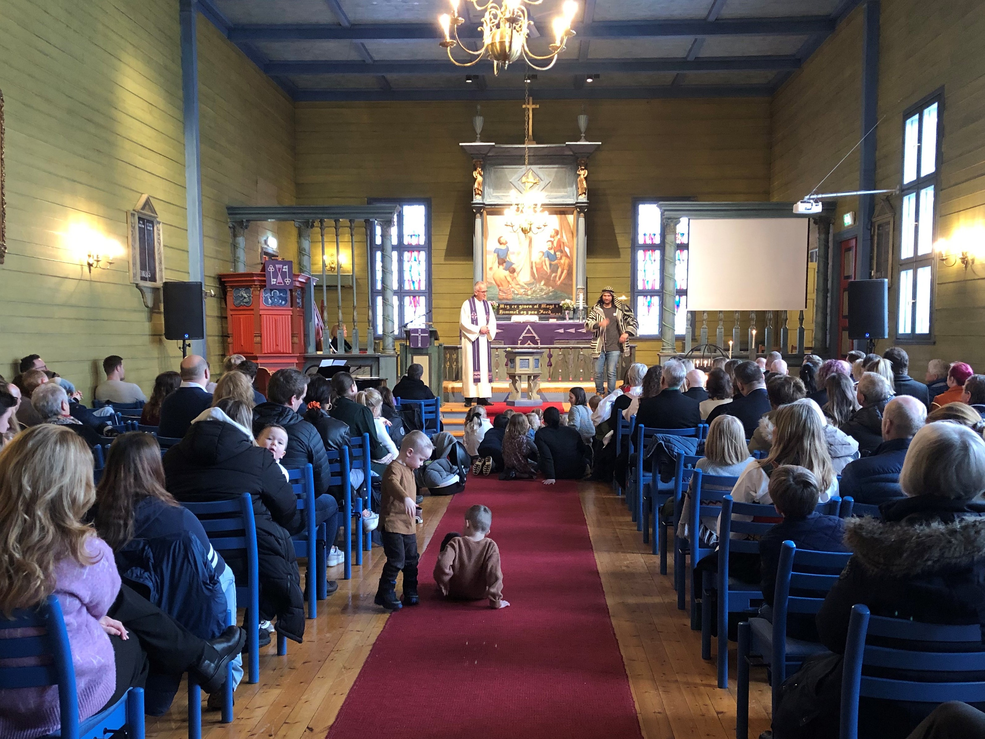 En prest og pedagog står framme i Blomvåg kyrkje, på gulvet framfor dei sit mange barn og høyrer på. Kyrkja er full av menneske som sit på stolane.