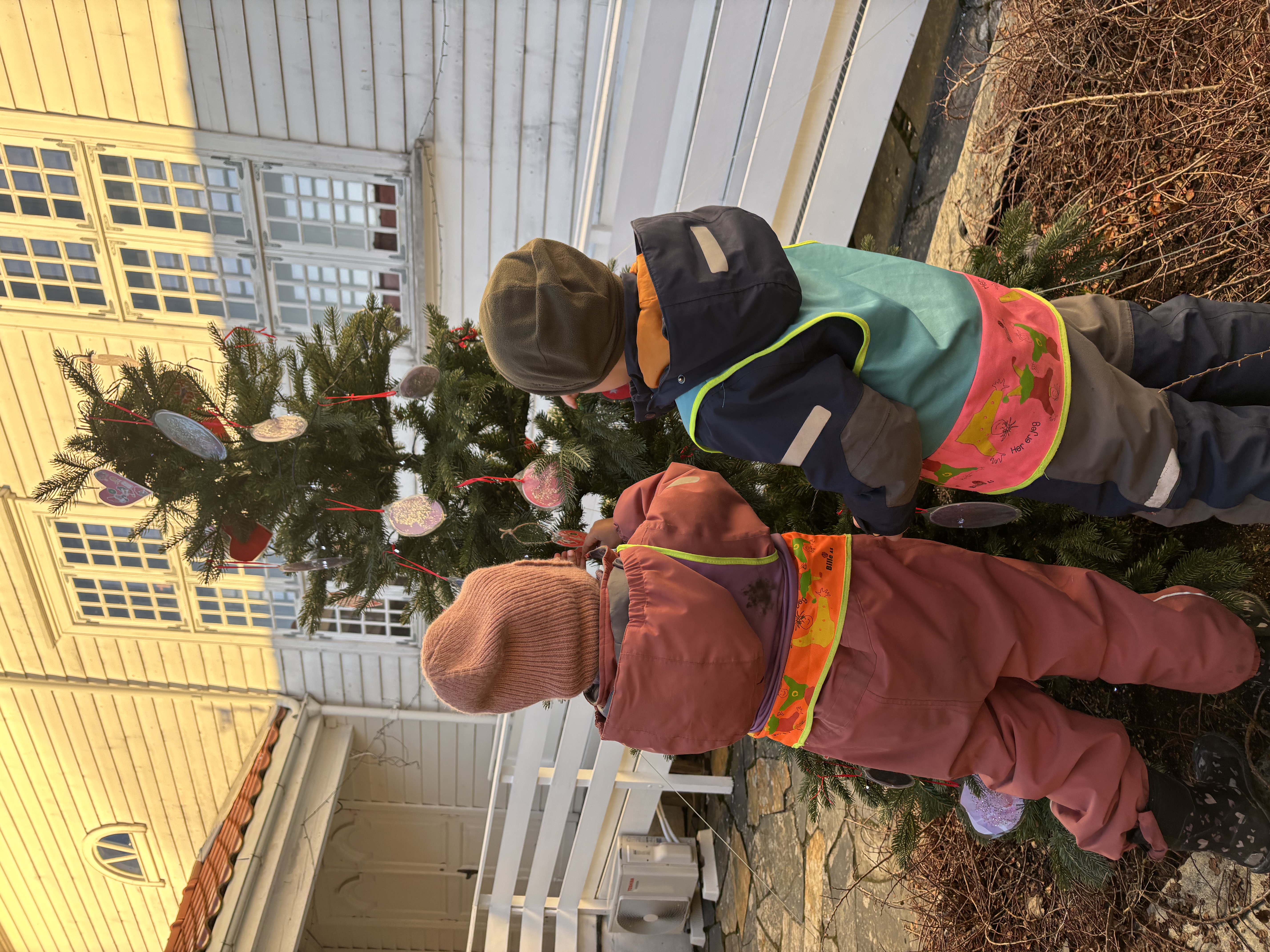 To barnehagebarn står i borggården og henger pynt opp et lite juletre