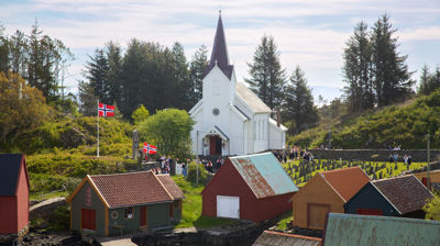 En hvit trekirke med flere naust framfor seg. Flere flagg er heist og man kan skimte mennesker framfor kirken