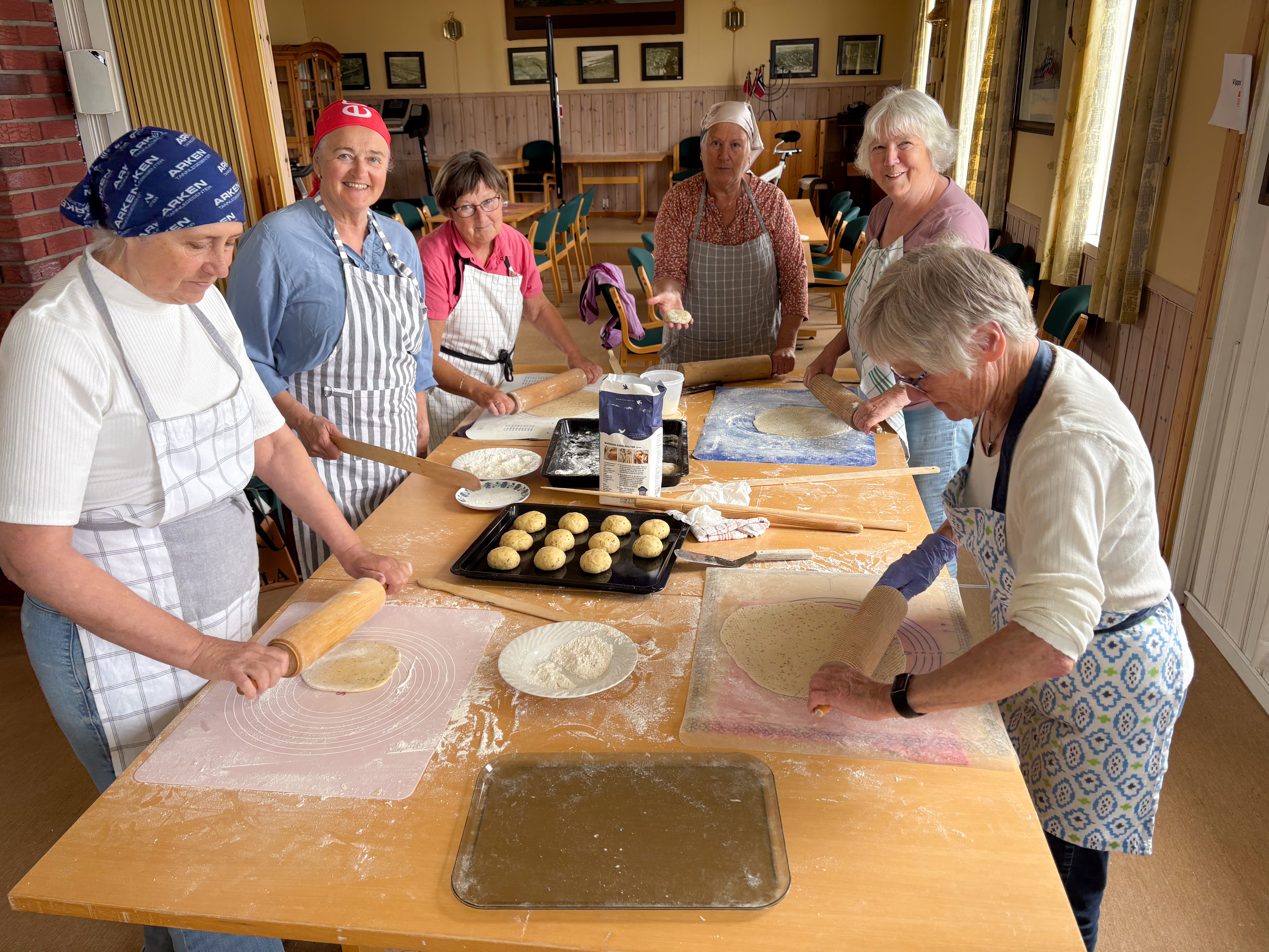 Fflere damer sår rundt et stort bord og baker og ruller ut lefser