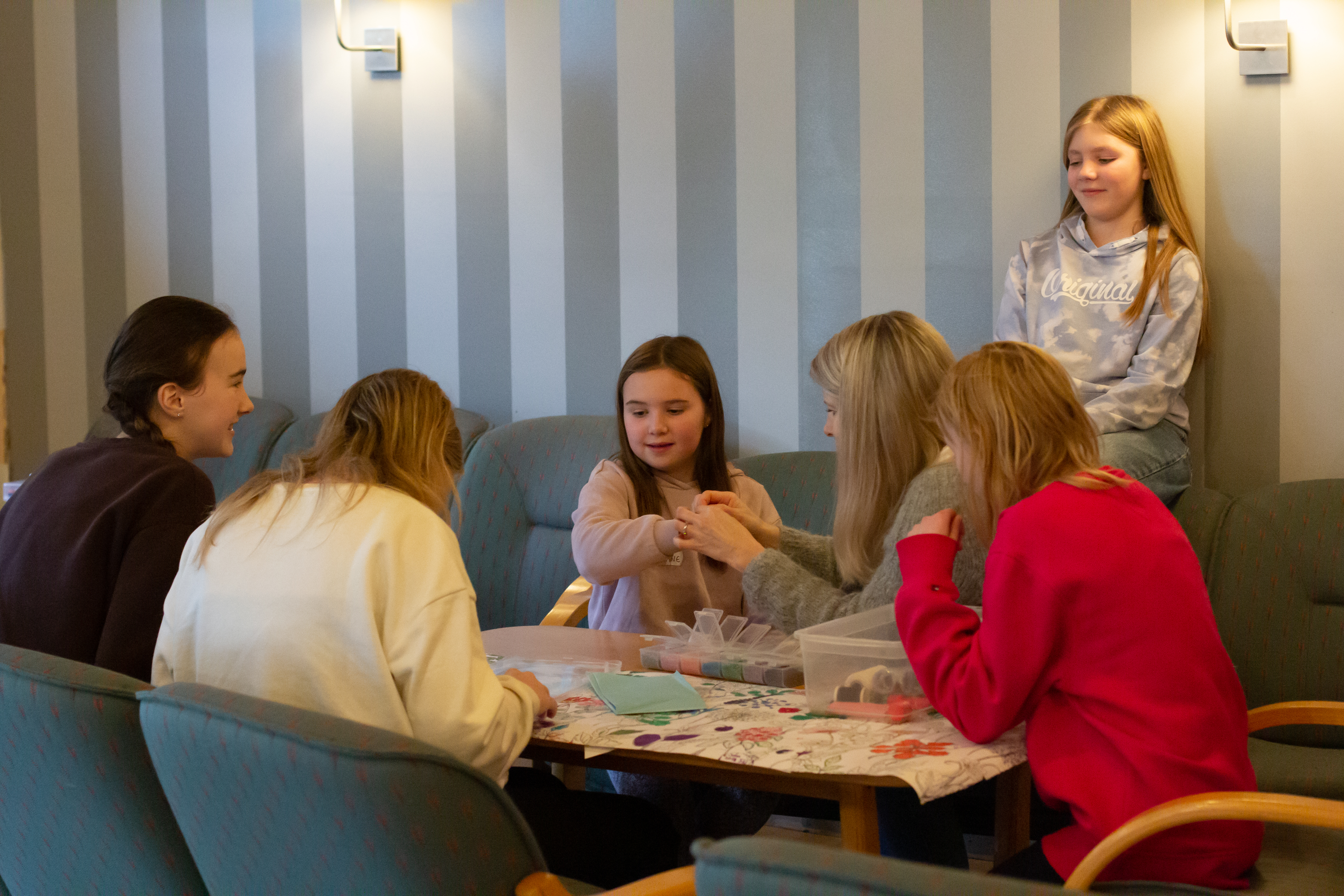 Barn og ungdommar sitt i ein sofagruppe og lagar armbånd