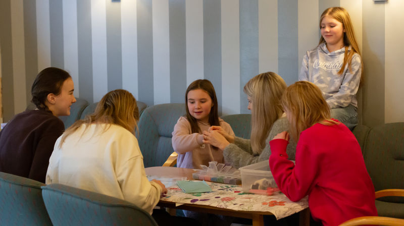 Barn og ungdommar sitt i ein sofagruppe og lagar armbånd