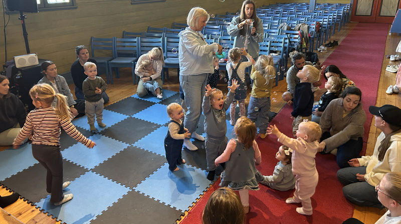 Små barn og voksne står og sitter på matter inni kirken, to damer holder maskiner som blåser ut såpebolber. barna koser seg og prøver å fange boblene