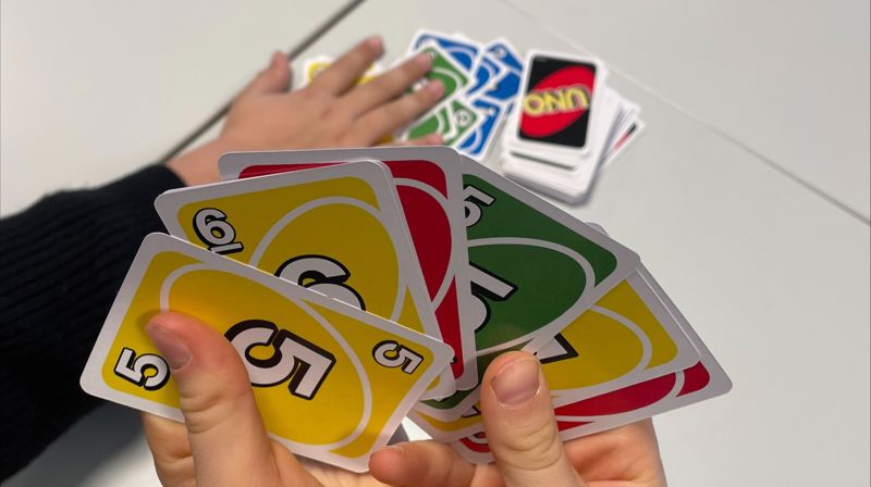 Et par hender holder opp uno-kort, en hånd i bakgrunnen trekker kort fra en haug