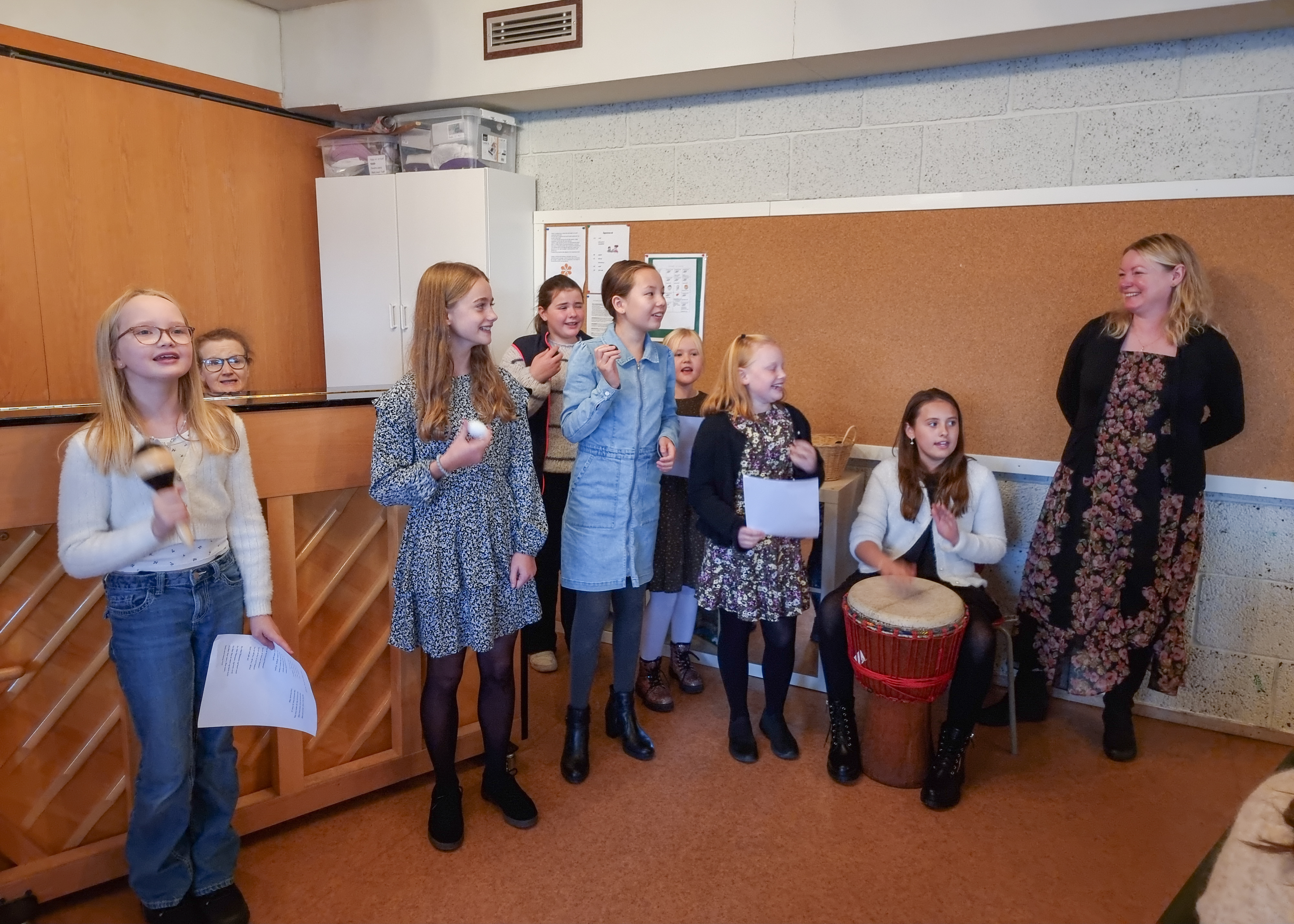 tweensjenter synger og spiller trommeinstrument, til høyre står en dame og er rørt