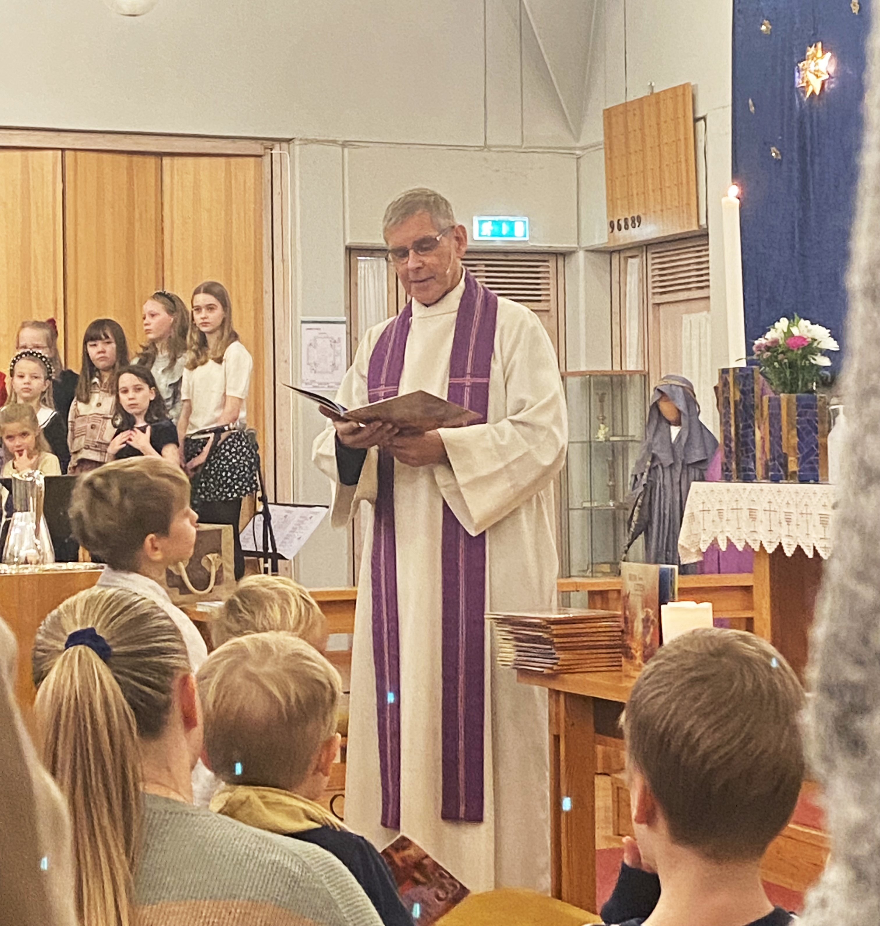 Presten står og leser fra juleboka med barn og voksne framfor seg