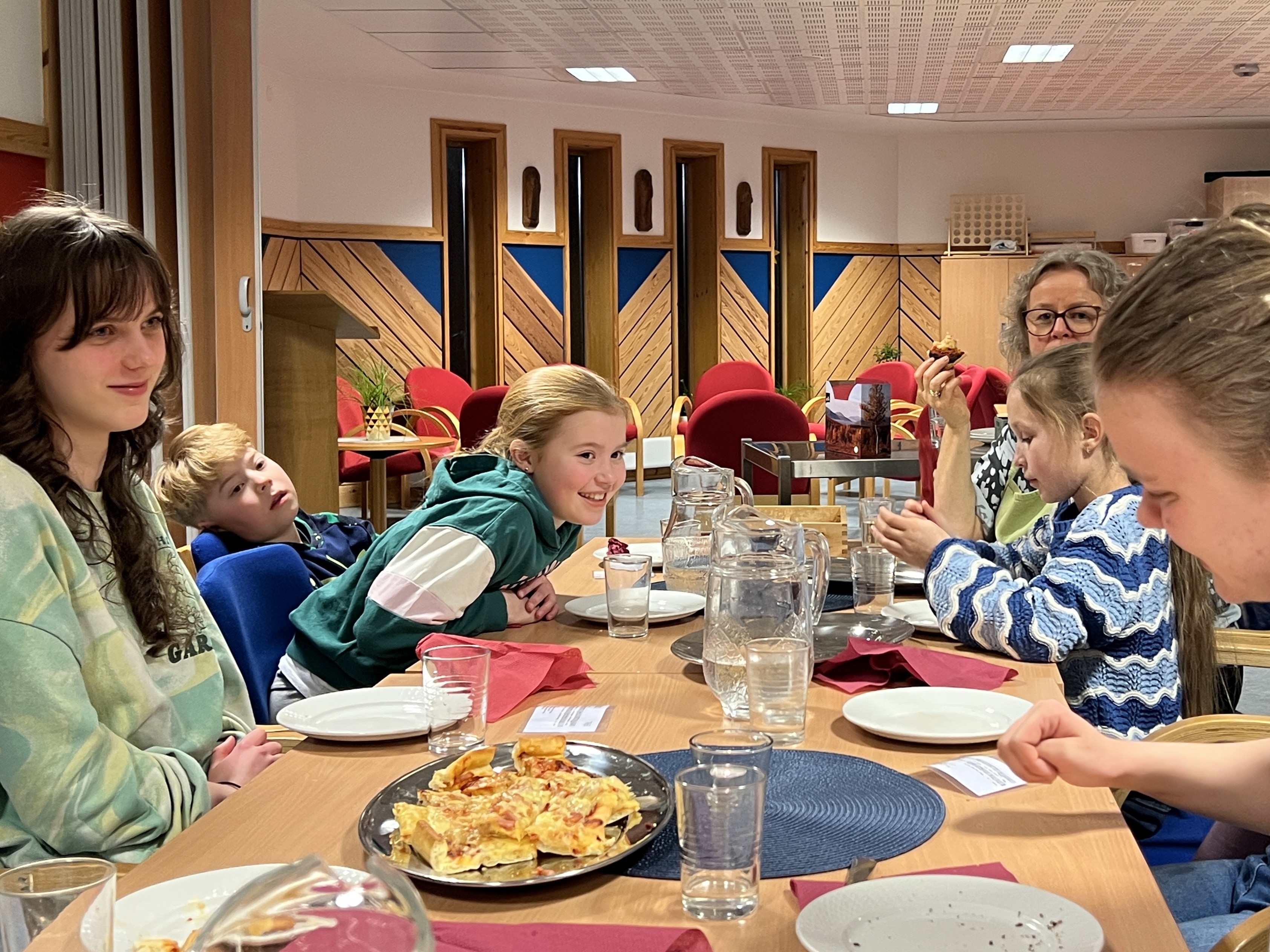 Barn og voksne sitter rundt et bord og spiser pizza, en jente lener seg over bordet og smiler