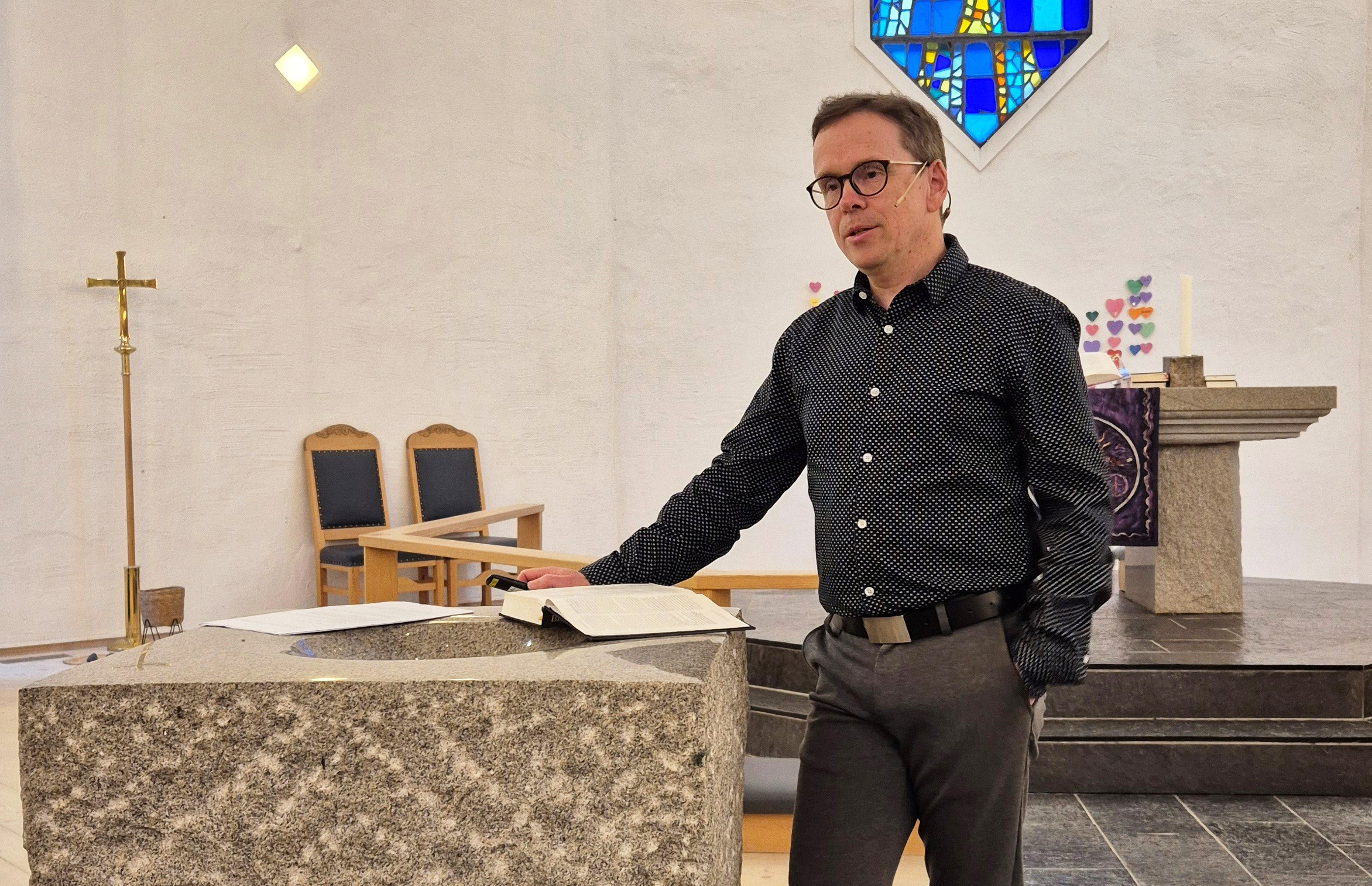 En mann står ved døpefonten i Sund kyrkje og snakker, han har en bok og et papir liggende på døpefonten. Han er kledd i mørk skjorte og grå bukse.