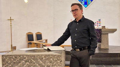 En mann står ved døpefonten i Sund kyrkje og snakker, han har en bok og et papir liggende på døpefonten. Han er kledd i mørk skjorte og grå bukse.